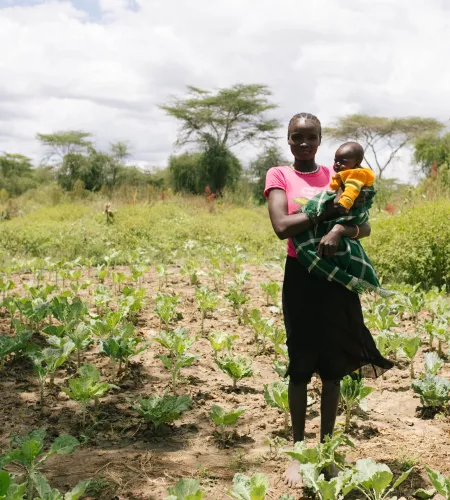 Weltflüchtlingstag: Frau mit Kind auf dem Arm in einem Gemüsebeet